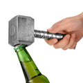 Hand using a hammer-shaped silver beer bottle opener to open a green beer bottle.